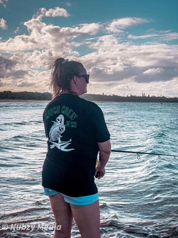 Rear view of female wearing Catch Crew N' Co t-shirt while fishing at the beach